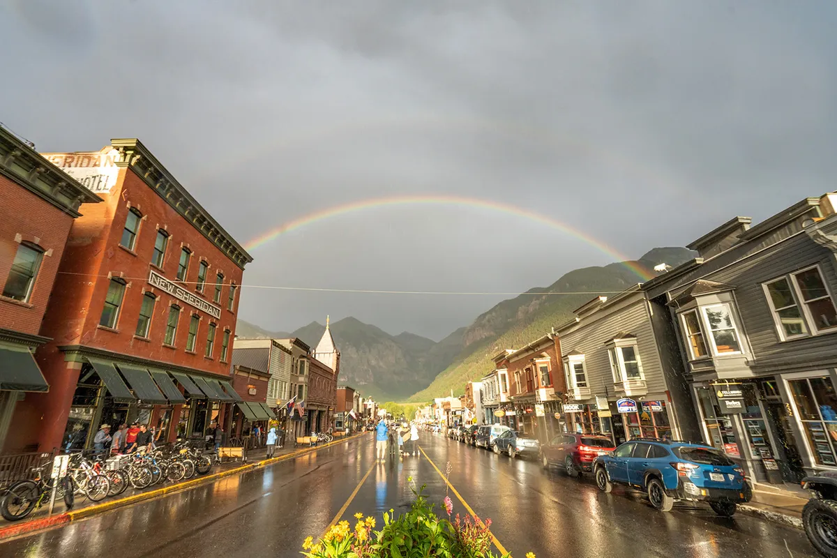 A vibrant, post-rain scene showcasing Telluride’s historic downtown with colorful buildings, a rainbow arching above wet pavement, and mountains in the background. People are seen walking, shopping, and dining, highlighting the town’s lively and welcoming atmosphere.