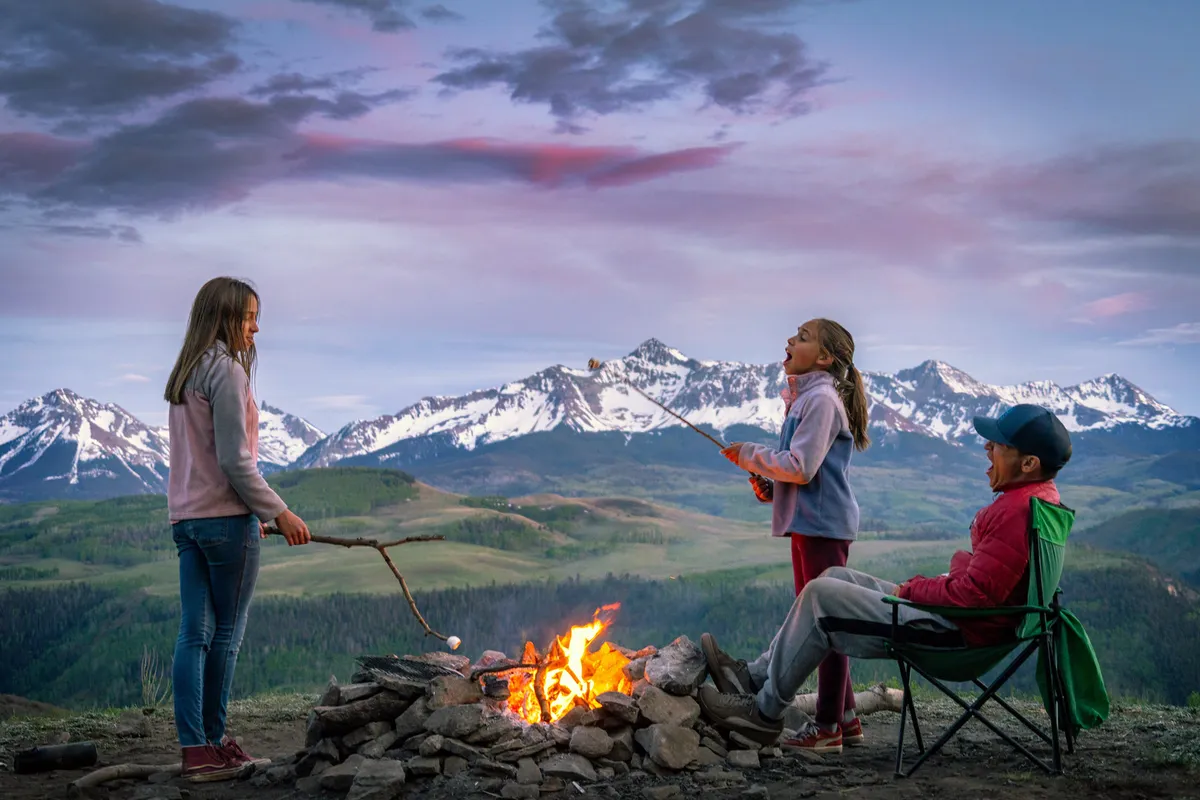 A joyful outdoor scene of children gathered around a campfire, roasting marshmallows, with an adult nearby and majestic mountains in the background. The setting evokes togetherness, nature, and lasting family memories.