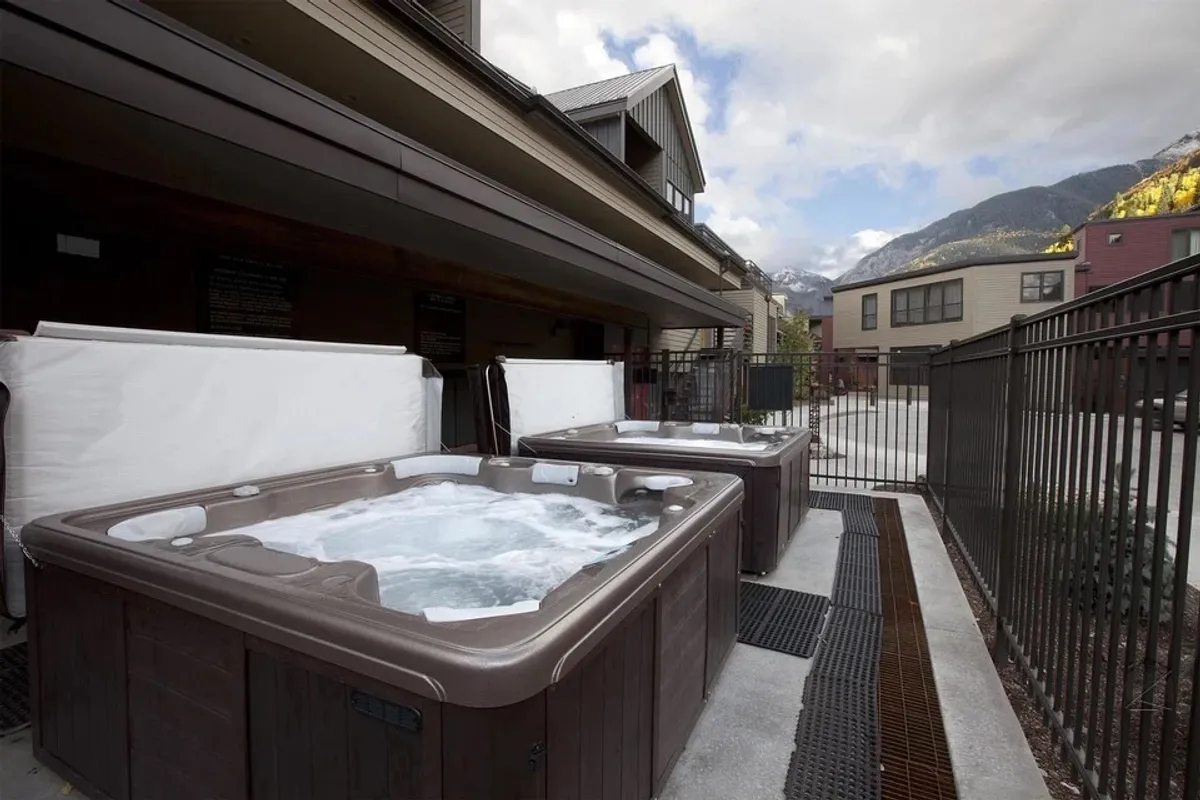 This image features steaming communal hot tubs framed by a stunning mountain vista and surrounded by lodge buildings. The inviting, bubbling water and close proximity to the condos embody the mountain-lodge lifestyle and relaxation opportunities available right outside the unit.