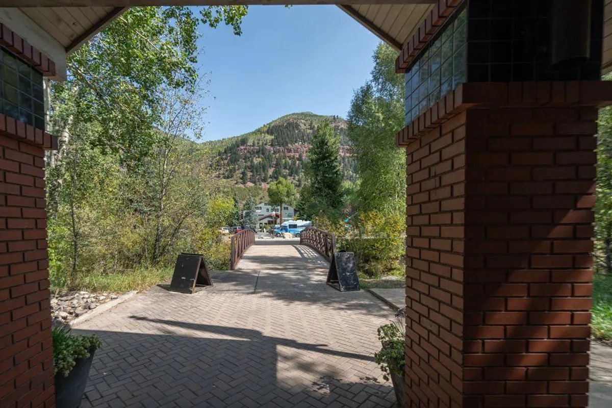 The image features a peaceful exterior scene with an arched entrance supported by brick pillars, a picturesque pathway, a bridge, and lush surrounding trees. The natural setting is tranquil, inviting families to enjoy outdoor walks and exploration.