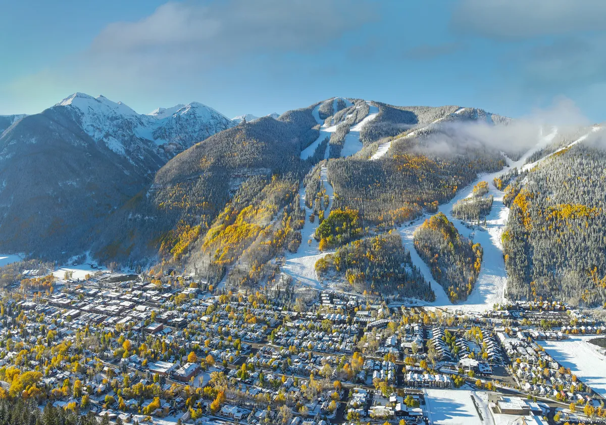 A panoramic view over Telluride featuring snow-covered mountains, ski slopes, colorful autumn trees, and the charming town below. The layered scenery conveys both natural beauty and proximity to local attractions.
