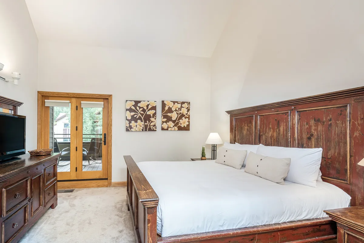 A relaxing bedroom scene with a comfortable bed, elegant wooden furniture, artwork on the wall, and sliding doors leading to a deck or outdoor space. The mood is tranquil, perfect for quiet mornings and restful nights.