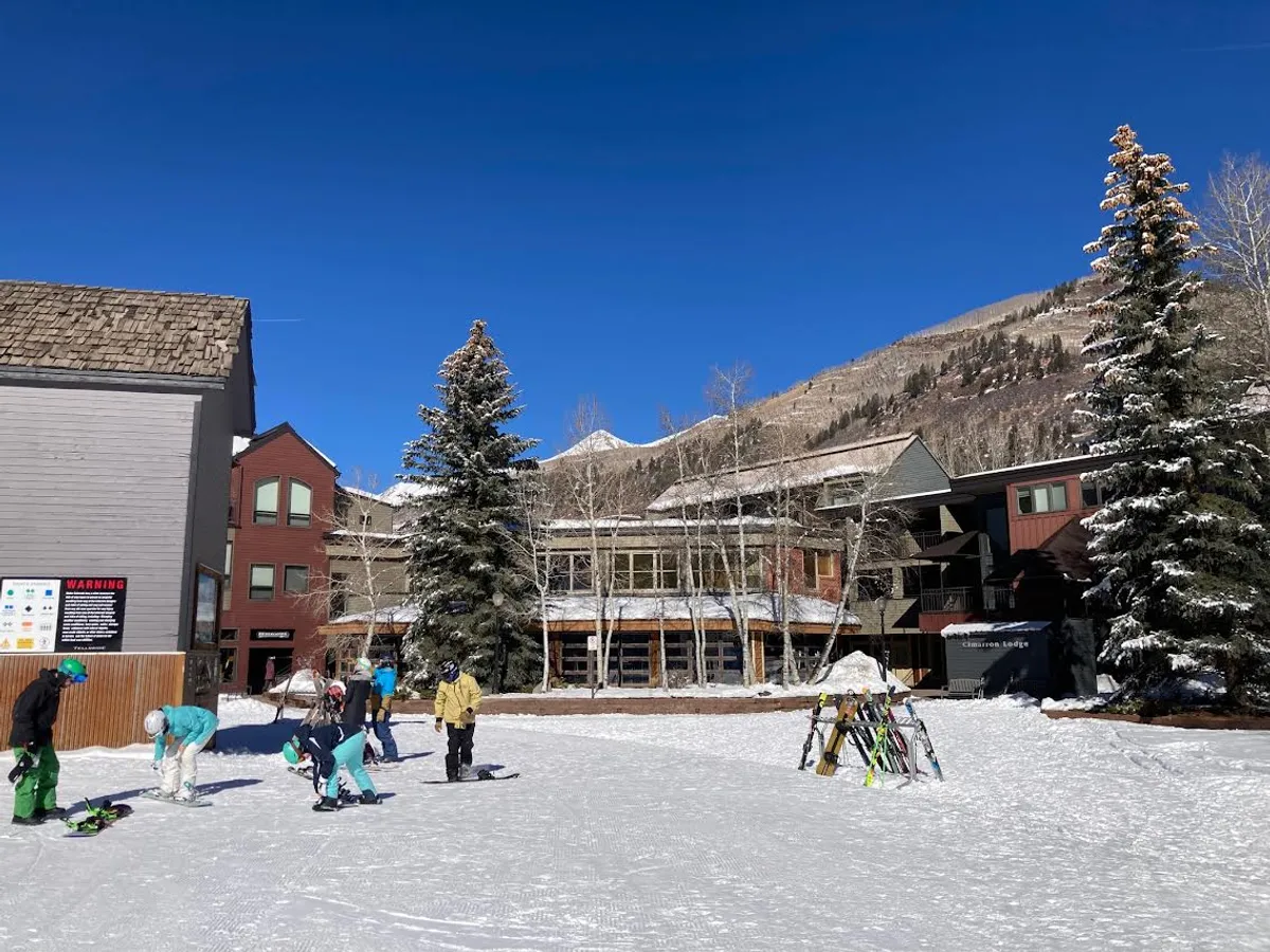 A vibrant winter scene featuring skiers gearing up at the base of the slopes, surrounded by trees, with the mountains rising in the background and lodge buildings visible nearby. The atmosphere is lively and full of anticipation for a day on the snow.