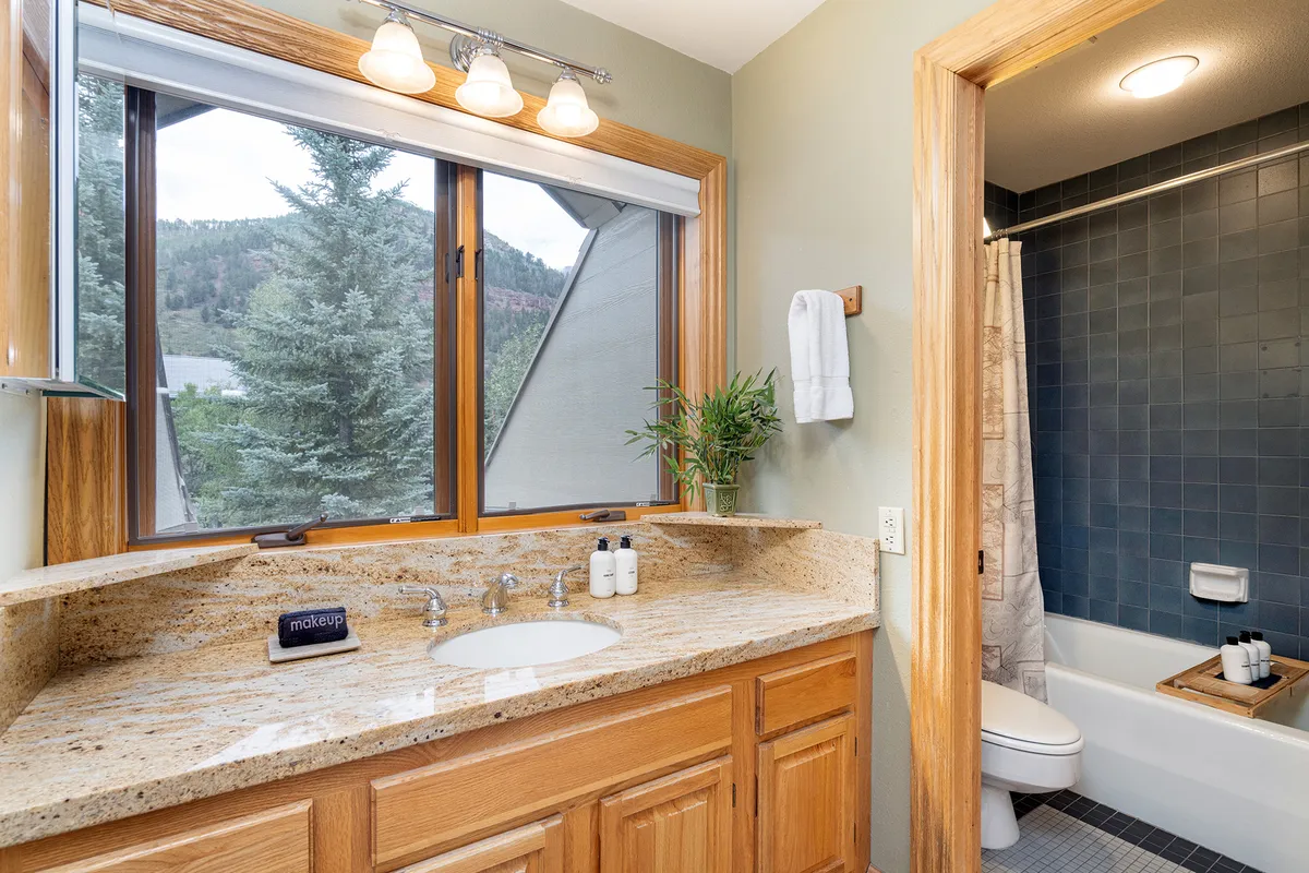 A bright, modern bathroom with a granite countertop, a deep soaking bathtub, a spacious shower area, and large windows letting in natural light. The wooden cabinetry adds warmth and elegance.