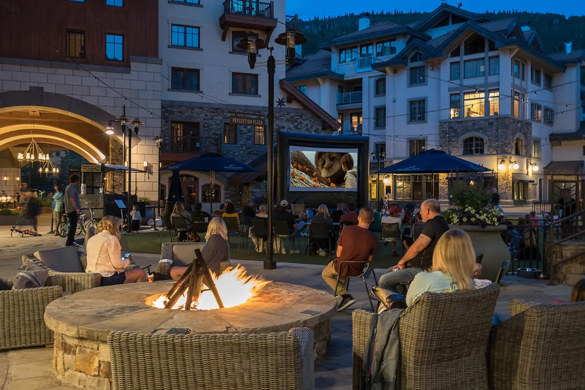 A group of people gathered around an outdoor fire pit, with a movie screen set up under the stars. The relaxed scene features warm firelight, cozy seating, and a backdrop of night sky, emphasizing communal enjoyment and relaxation.