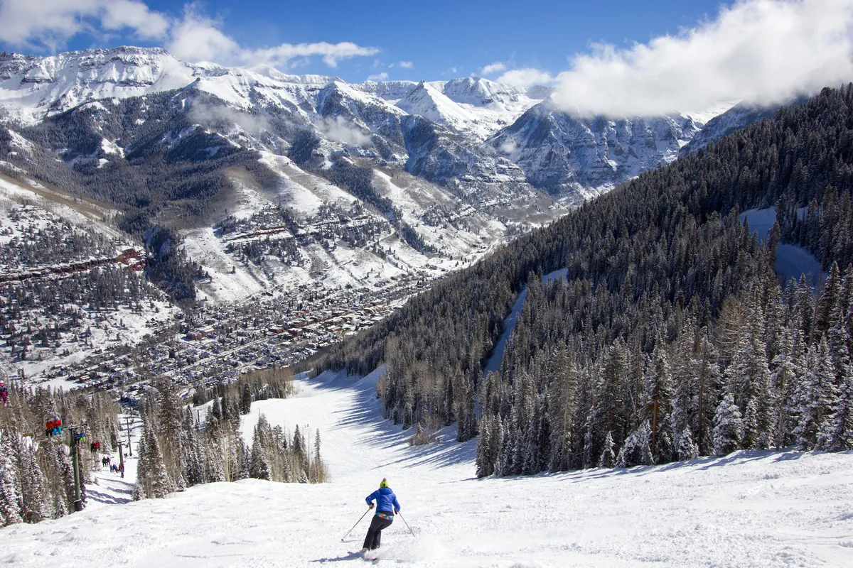 A breathtaking view of snow-covered mountains with a ski slope, a skier in the foreground, and the town nestled below. The scene is vibrant and active, capturing the adventure of skiing and the picturesque setting of Telluride.