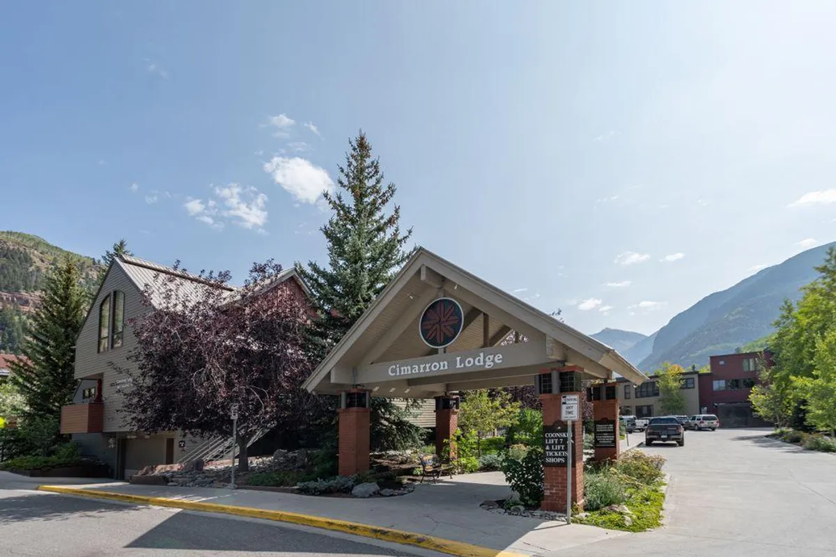An exterior view of the Cimarron Lodge entrance, featuring the lodge sign, welcoming awning, lush trees, and mountain backdrop. The setting feels inviting and hints at the easy walkability to shops, dining, and the gondola as described in the section.