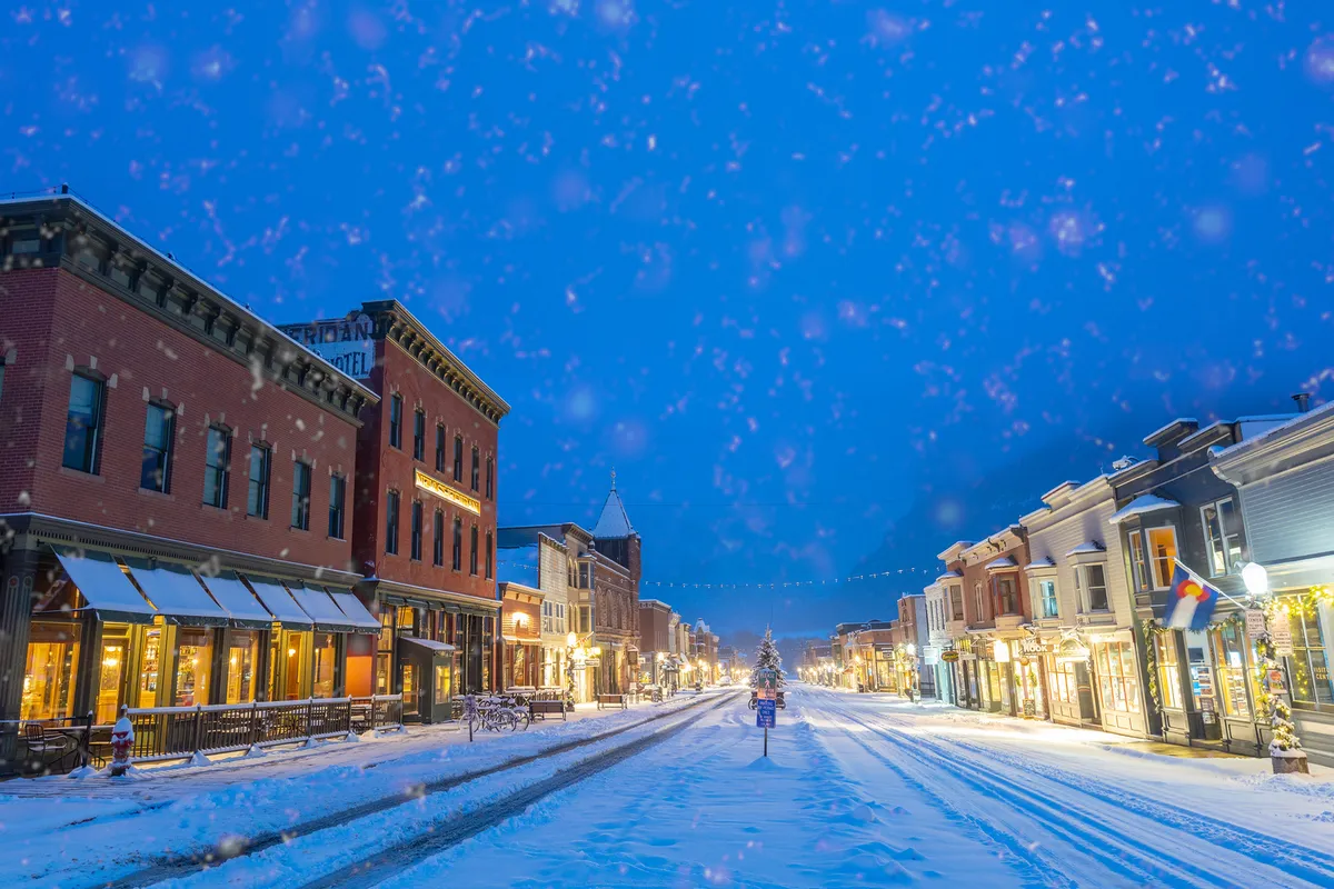 A snow-covered street lined with historic buildings, festive decorated trees, and inviting storefronts. The atmosphere hints at winter festivities, community gatherings, and the cozy charm of Telluride's downtown area after a day on the slopes.