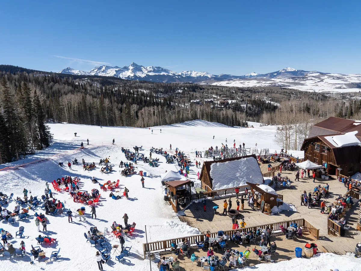 A vibrant scene at a ski lodge with snow-covered slopes, a crowd of people enjoying outdoor seating, and a stunning mountain range in the background. The colorful chairs and social atmosphere suggest communal moments after a day of activity.