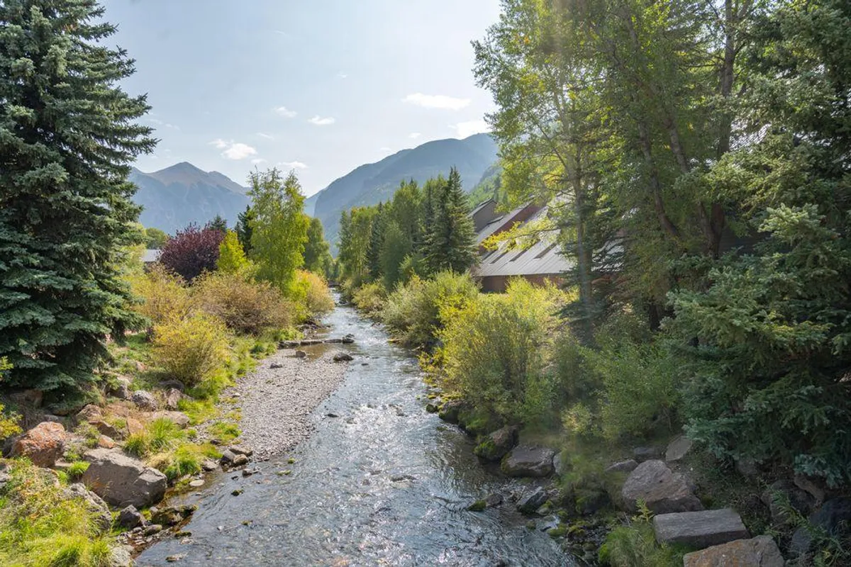 A tranquil summer view of a flowing river bordered by lush trees, with a mountain backdrop. A building is partially visible, hinting at the proximity of the lodge to natural beauty. The peaceful scene suggests access to the River Trail for hiking and biking.